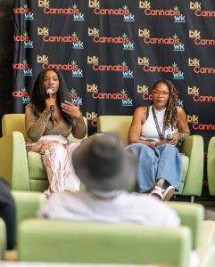 Two women sit on stage at a conference, speaking into microphones. Behind them is a backdrop with 'Black Cannabis WK' logos and text.