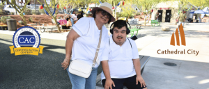 Woman wearing a hat and sunglasses stands next to a seated young man with headphones in a sunny outdoor park setting.