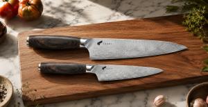aski 8-inch chef knife and 5.5-inch utility knife with Damascus steel blades and dark wood handles resting on a wooden cutting board alongside fresh tomatoes and garlic.