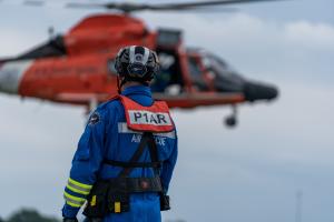 P1AR instructor conducting live flight training; image shows P1AR instructor standing in foreground with MH65 helicopter in the background.