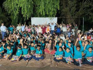 Large group of students, families, and community members gathered outdoors at night, smiling and raising their hands during a joyful community celebration, with children seated in the front holding gift packages.