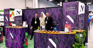 Three members of the New Acai Amazonas team standing at the company's purple-branded trade show booth displaying organic acai and superfruit powder samples in wooden bowls.