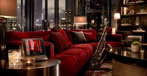 Elegant lounge with burgundy velvet seating, nighttime city skyline, saxophone, lemon water, and “Plan of Perfection” album artwork.