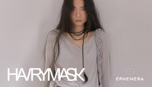 Model wearing a HAIRYMASK braided grey scarf and black braided cord necklace with hair charm, from the Seoul accessories brand's lookbook — launching in the U.S. through EPHEMERA.