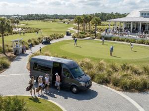 An aerial view of a dark Mercedes Sprinter van marked Tampa Luxe parked at a golf clubhouse as four guests unload golf bags beside a putting green.