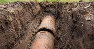 Exposed clay sewer pipe with a separated joint in a damp residential utility trench, viewed from above with the lawn softly blurred in the background.