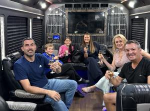 About sixt adults seated together inside a party bus with leather lounge seating and lit ceiling panels, smiling toward the camera.