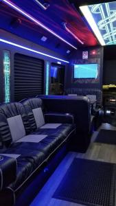 The interior of a party bus, empty, showing black and grey leather lounge seating, wood flooring, a wall-mounted screen and blue and teal LED lighting.
