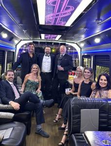 Eight adults in formal wear seated and standing inside a party bus with blue and pink LED ceiling lighting.