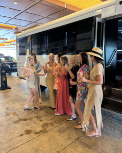 Eight women in resort wear holding champagne flutes, standing in a line beside a black and cream motorcoach under a covered entrance.
