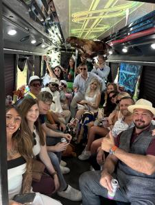 A full party bus of about sixteen adults in derby attire, seated and standing under a lit ceiling panel and a horse decoration.