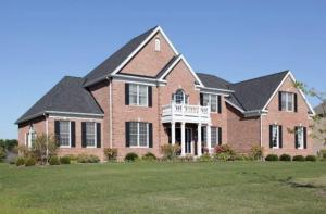 Large two-story brick house with multiple peaked roofs, black shutters, a white balcony, and a spacious green front lawn under a clear blue sky