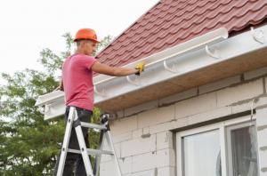 A construction worker installing a white rain gutter along the edge of a residential roof while standing on a ladder.