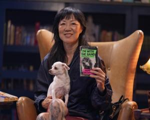 Margaret Cho poses in a leather library chair, holding her dog and her personal copy of Pamela Des Barres' memoir I'm With the Band.