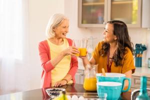 An elderly woman and a female caregiver smiling and sharing glasses of orange juice together in a bright kitchen.