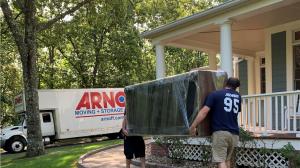 Two uniformed Arnoff employees carrying a large, wrapped piece of furniture down a walkway towards a white Arnoff Moving & Storage truck parked in a residential driveway.  Image 3 (image_2.png)