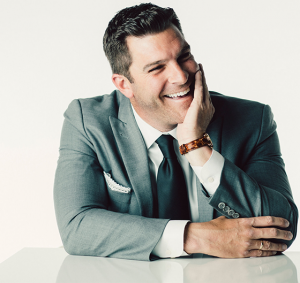 Portrait of Garrett Weston in a grey suit and dark tie, seated and smiling with his hand against his cheek.