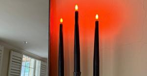Celestial At Home rechargeable black flameless Halloween wax taper candles with orange glowing flame tips, standing in twisted iron candlesticks against an orange-washed wall beside a mirror