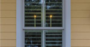 Celestial At Home battery operated window candles lit at matched height in a shuttered double-hung porch window at dusk, seen from the street