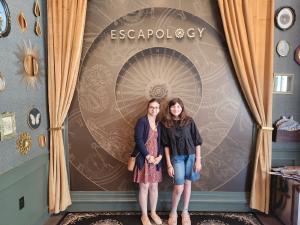 Two young women standing together and smiling in front of an escape room entrance with a decorative circular design and gold curtains.