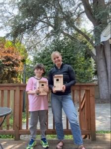 An adult woman and a young boy standing on an outdoor deck, each holding a wooden birdhouse they built together.