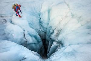 Rescuer in red technical gear approaching a deep crevasse on a glacier.