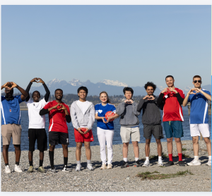 Shekinah Grace Moyes with a diverse group of performers in sportswear standing by the Vancouver waterfront, forming heart shapes with their hands, with mountains in the background.