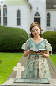Shekinah Grace Moyes holds an open Bible outside a church, wearing an elegant green dress, with a Christian “Joined in Love” cross in the foreground.