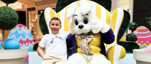 A boy sitting next to a person in a white Easter bunny costume on a large yellow and white striped chair, surrounded by Easter decorations.