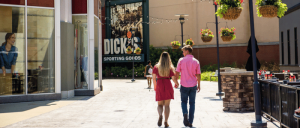 Couple walking hand in hand along a sunny outdoor shopping area with hanging flower baskets and a Dick’s Sporting Goods store.