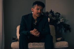 Independent alt-pop musician Danny Silvertone wearing a dark blazer and cross earring in a dimly lit setting.