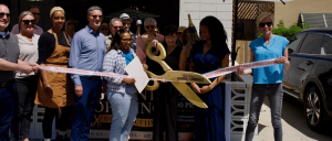 Group of people celebrating a ribbon-cutting ceremony outside, one woman holding large gold scissors cutting the ribbon.