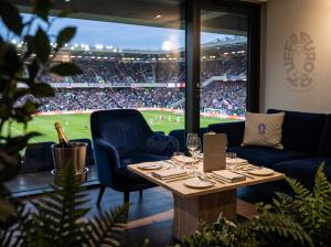 An elegant lounge-style hospitality suite in a stadium, dressed with fine furniture and branded with EURO 2028 logos, showing indicative view into stadium bowl