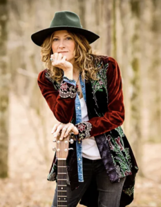 A outdoor portrait of musician Michelle Malone looking thoughtful and resting her chin on her hand, leaning on the neck of an upright acoustic guitar. Attire & Styling: She wears a wide-brimmed dark green felt hat over long, wavy light brown hair, a whit