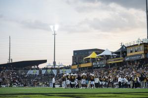 Kennesaw State Owls football players gather on the sideline at a packed stadium, surrounded by fans in KSU’s black and gold.