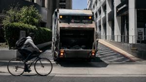 A bicyclist passes in front of a garbage truck backing out of a commercial driveway, illustrating the blind-spot and visibility risks that can lead to serious garbage-truck crashes involving cyclists and pedestrians.