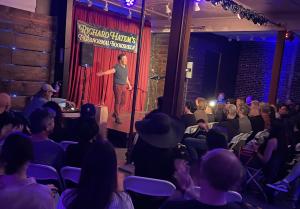 A performer on stage with open arms welcoming a seated audience in an intimate venue under bright stage lighting, with a banner reading "Richard Hatem's Paranormal Bookshelf" hanging on red backdrop curtains behind him.