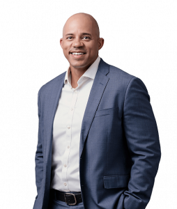 Jordan Brice standing facing forward against a plain white background, wearing a light blue suit jacket with matching trousers, a white dress shirt, and a dark belt. They are clean‑shaven, smiling, and posed in a confident, professional manner.