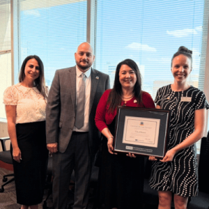 Four professionals standing in an office, one holding a framed certificate, all smiling at the camera.