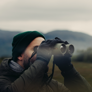 A man in a green beanie looks through Avilume binoculars, gloved hands raised to his eyes, with mountains blurred in the background.