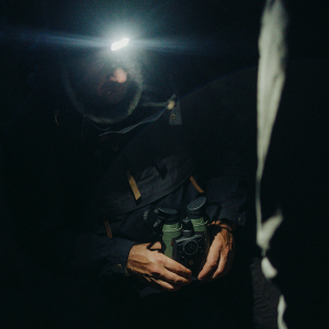 A man at night wears a headlamp and holds olive-green Avilume binoculars close to his chest; another person stands beside him in the dark.