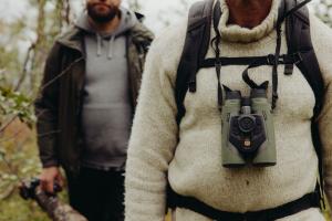 A man in a cream wool sweater wears olive-green Avilume binoculars around his neck; a second hiker stands blurred in the woods behind him.