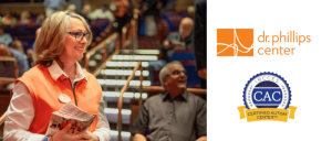 Woman in an orange vest holding pamphlets smiles in a theater, alongside the Dr. Phillips Center and Certified Autism Center logos.
