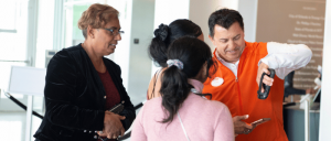 A man in an orange vest assists three women with scanning their phones in a bright, modern indoor setting.