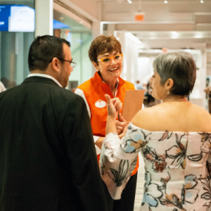 Three people are engaged in a cheerful conversation in a bright indoor setting, with one woman wearing an orange vest and glasses.