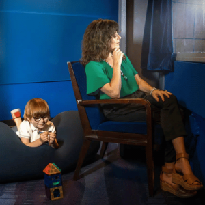 A woman in a green top sitting on a chair looking out a window, while a boy with glasses lies on a beanbag nearby.
