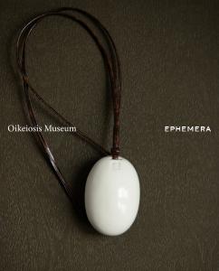The OIKEIOSIS MUSEUM Eggmic in White, an oval hand-formed ceramic pendant on a twisted leather cord, on an olive textured ground.