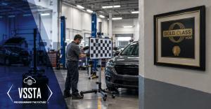 A modern auto repair shop with a technician performing an ADAS calibration on a vehicle using a calibration target. The facility features professional automotive equipment and a framed “Gold Class” I-CAR certification plaque on the wall, with VSSTA brandi