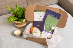 Open WeCARE™ Loss Support Kit box containing a purple and green guide, note cards, a journal, and comfort items on a table next to a candle and plant.