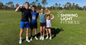 Four homeschool children of different ages smiling and flexing their arms on a grassy field beside the Shining Light Fitness logo.